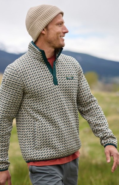 A person in a patterned pullover and beanie stands outdoors with mountains in the background.