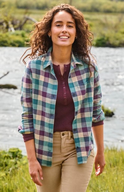 Woman with curly hair in plaid shirt and beige pants stands outdoors near a river with greenery in the background.