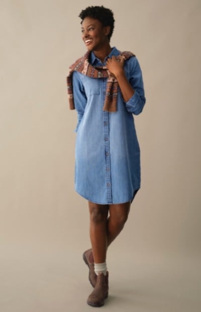 A woman in blue denim dress, brown boots, and scarf stands with legs crossed against a plain background.