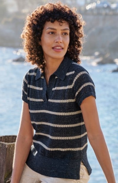 A womab with curly hair wearing a short-sleeve, striped knit shirt sits outdoors near water and rocks.