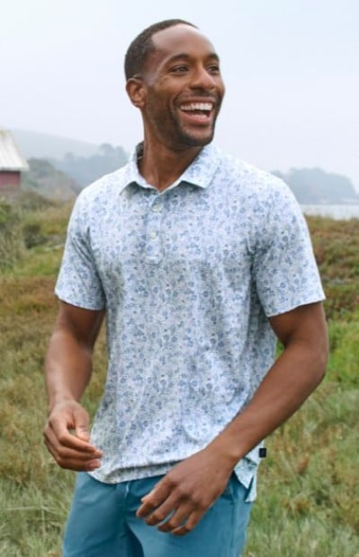 Person wearing a light patterned polo shirt and blue shorts while standing outdoors in a grassy landscape.