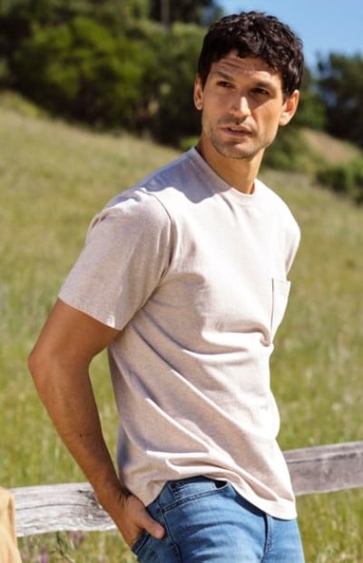 Person in a light T-shirt and jeans standing beside a wooden fence in a field.