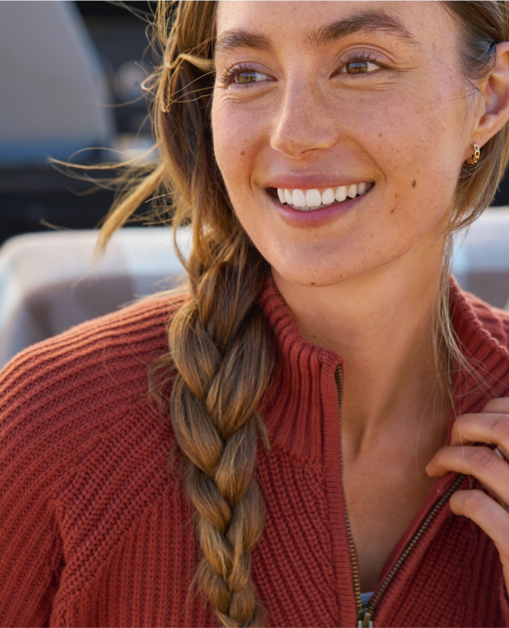 Woman in a rust-colored sweater and braid over shoulder sits outdoors.