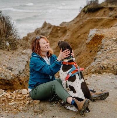 A woman sitting on the ground by water petting a dog.