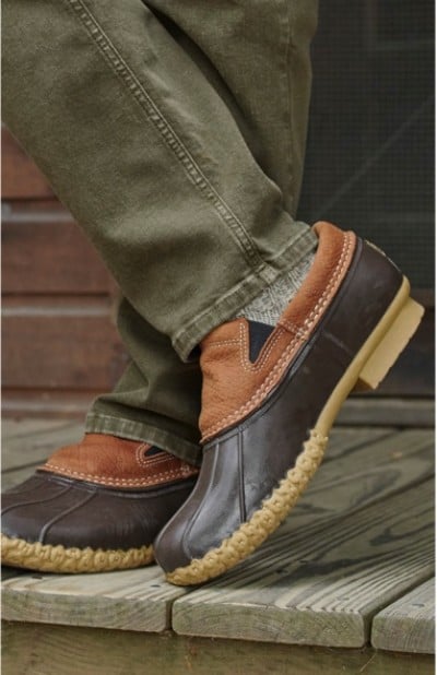 A person wearing brown and tan duck boots with green pants stands on a wooden deck.