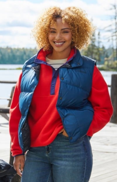 A woman with curly hair wearing a red sweater, blue puffer vest, and jeans stands outdoors near a lake.
