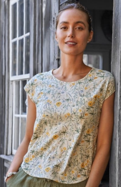 A woman wearing a floral patterned shirt and green pants stands against a wooden wall with windows.