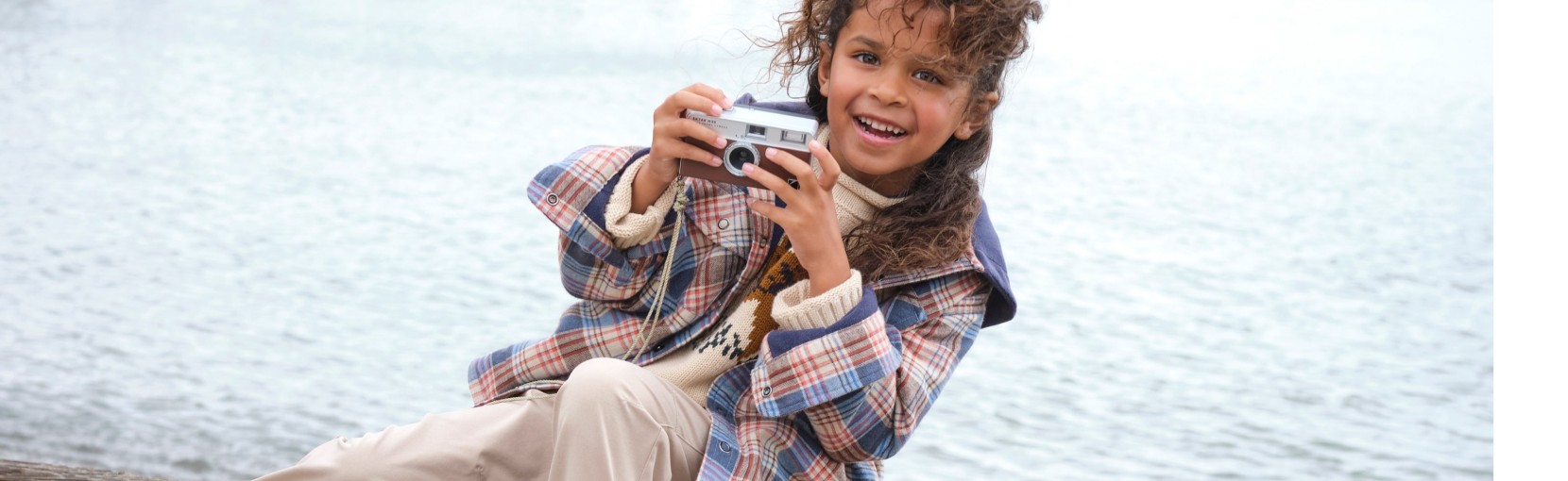 A child sitting outdoors near water, holding a small camera, wearing a plaid jacket and light-colored pants.