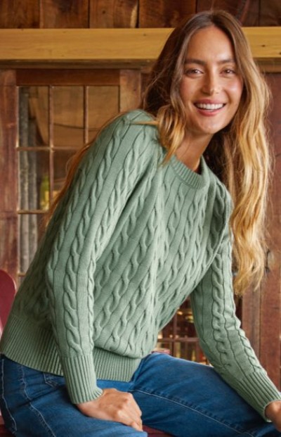 Woman wearing a mint cable‑knit sweater and blue jeans, seated indoors in a rustic wooden setting.