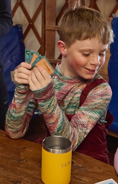 A boy in colorful L. L. Bean winter gear holds playing cards at a wooden table with a yellow L. L. Bean cup.