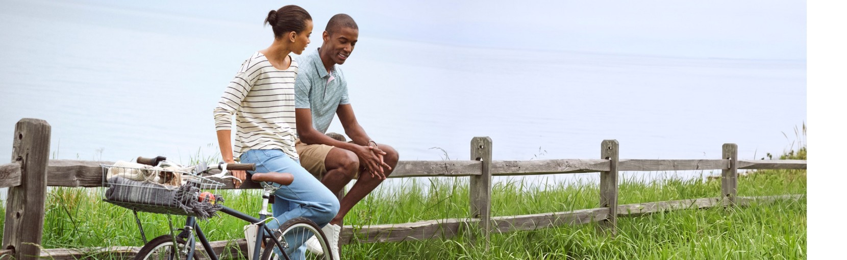 Two friends sit on a rustic wooden fence beside a bicycle, overlooking a grassy field.