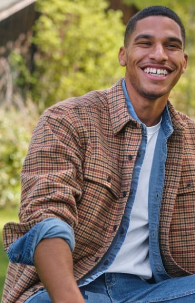 A man sits outdoors, wearing a plaid shirt and jeans, with a lush forest in the background.