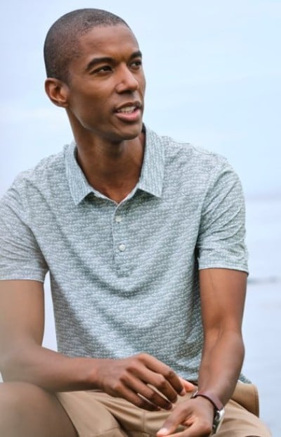 Person seated outdoors in a gray patterned polo shirt and shorts with a clear sky in the background.