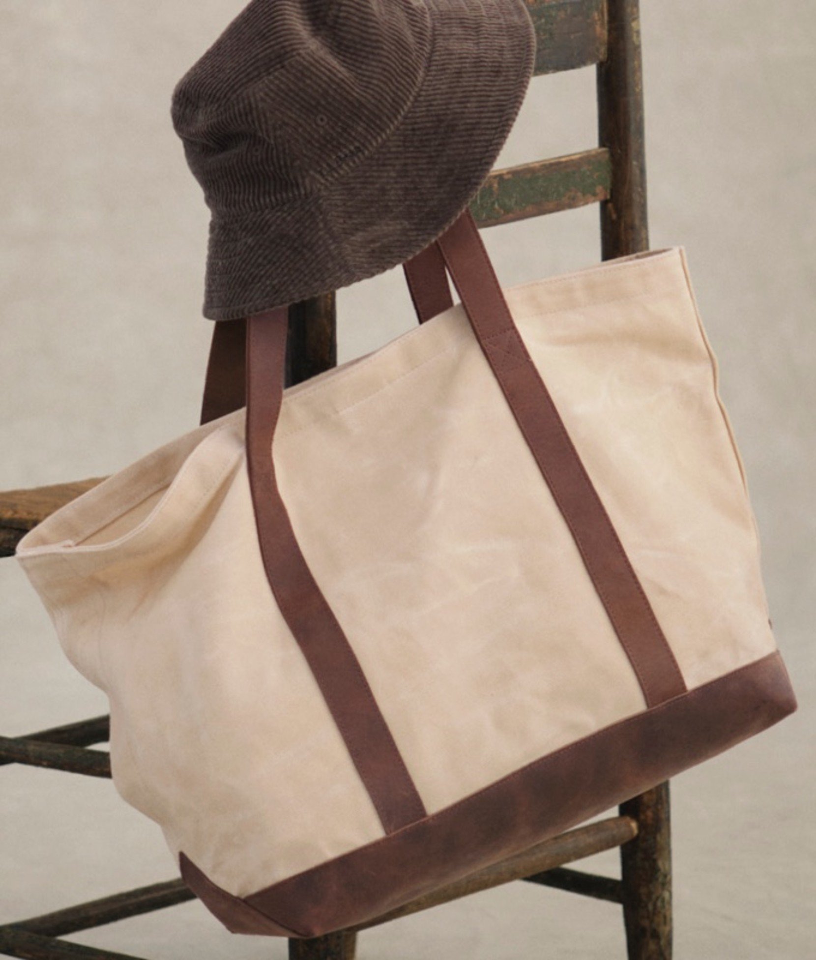 Beige Boat and Tote with brown straps and corduroy hat on wooden chair. 