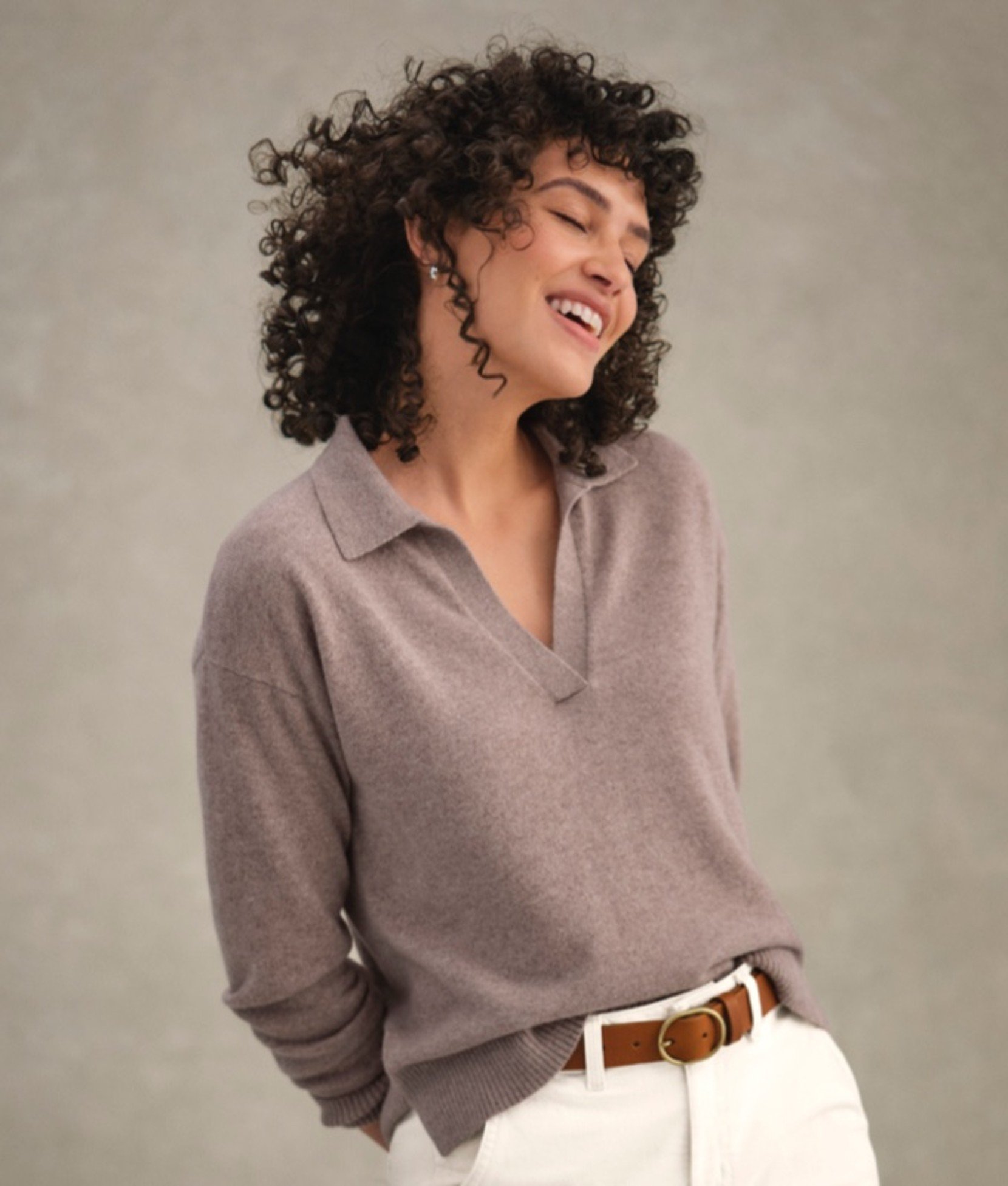 A woman in a mauve collared sweater tucked into white pants, standing.