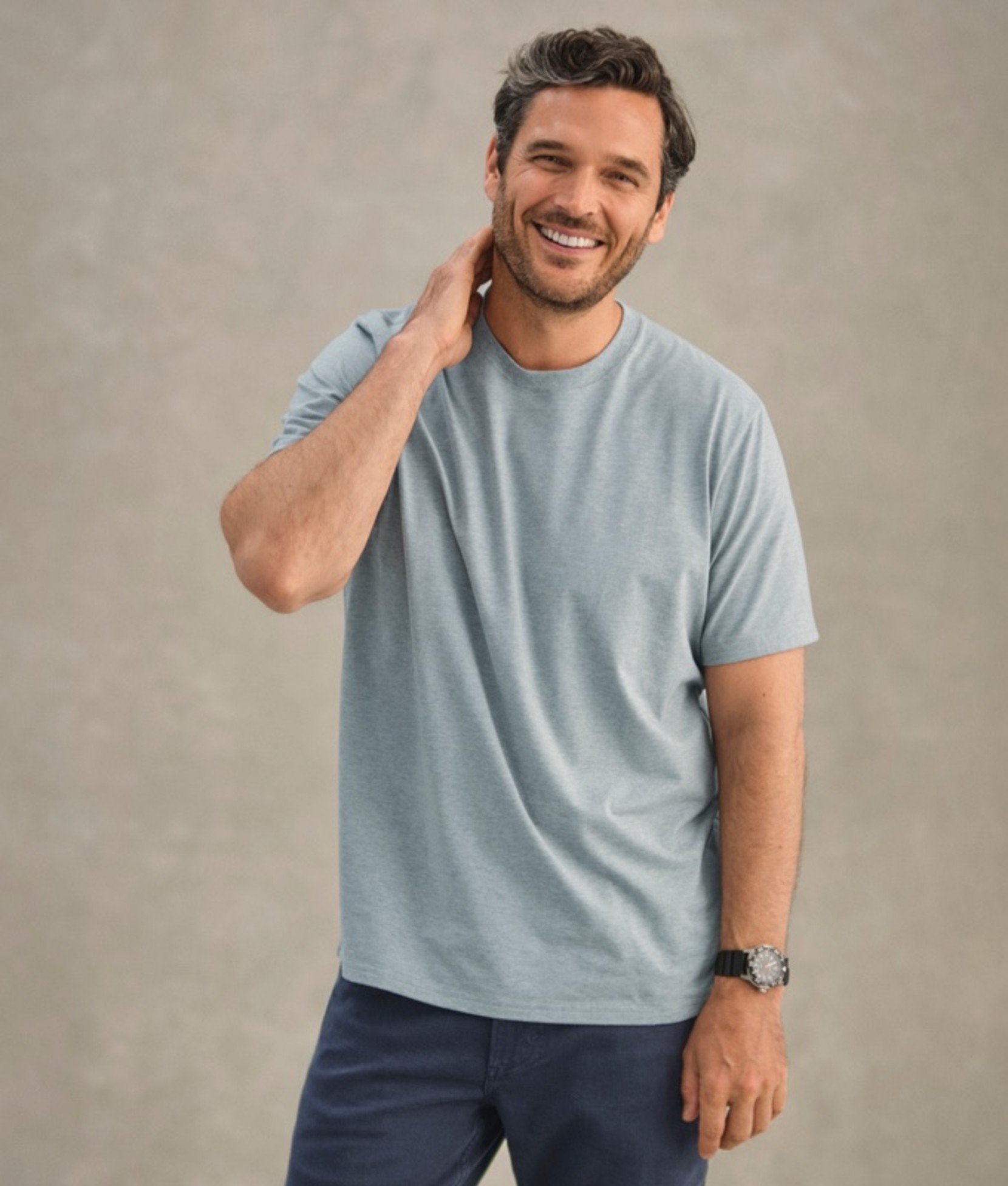 A man in light gray t-shirt and dark pants, touching neck with hand.