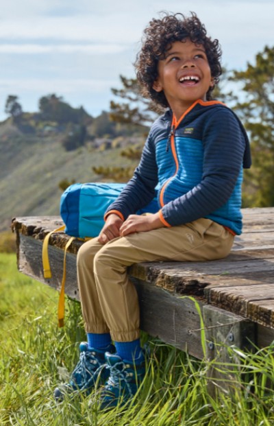 A young boy sits on a bench outdoors with a L L Bean backpack beside them, surrounded by nature.