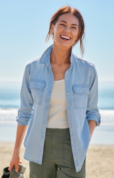 Person in light blue button-up shirt over white top and green pants stands on a beach with the ocean in the background.