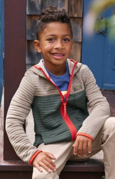 Child wearing a colorblock zip-up jacket with red accents over a blue shirt and beige pants, seated on outdoor wooden steps.