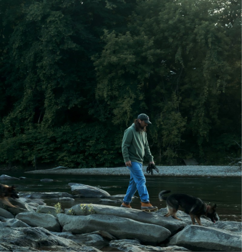 Noah Kahan wearing a green hoodie and L. L. Bean Boots walks on river rocks with his two dogs.