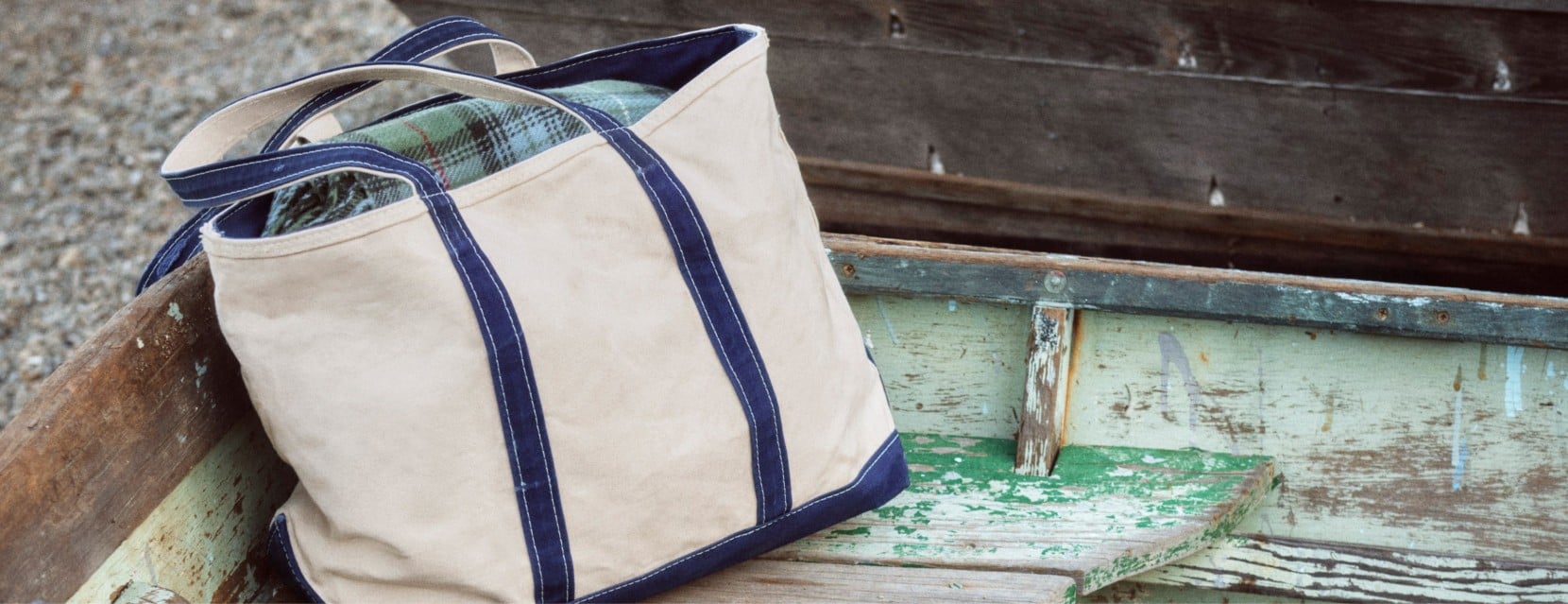 Canvas tote bag resting inside a weathered wooden boat, with a plaid shirt peeking from the top.