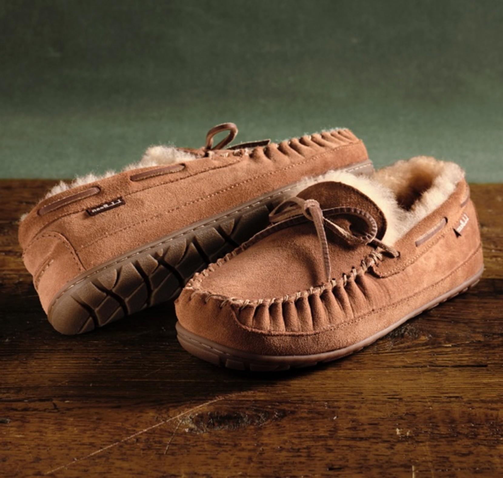 Tan suede moccasin slippers with fleece lining and bow detail on a wooden surface.