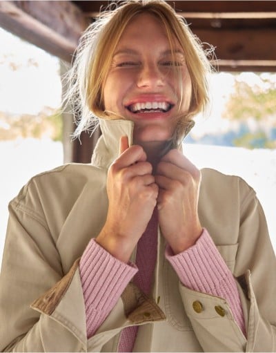 Person adjusts collar of a beige trench coat layered over a pink ribbed sweater.