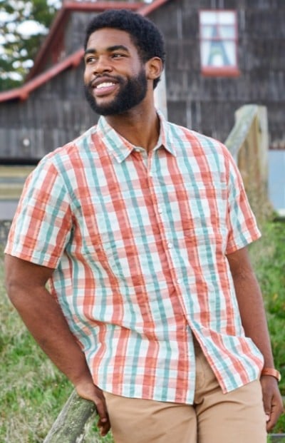 A man wearing a red, white, and blue plaid shirt and beige pants is leaning against a wooden fence.