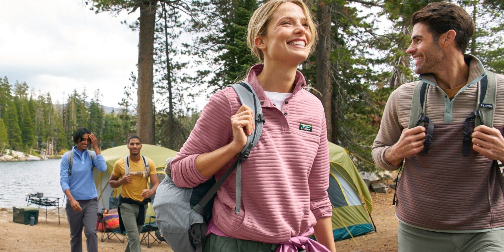 Group of hikers with backpacks walking near lakeside tents in a forest campsite.
