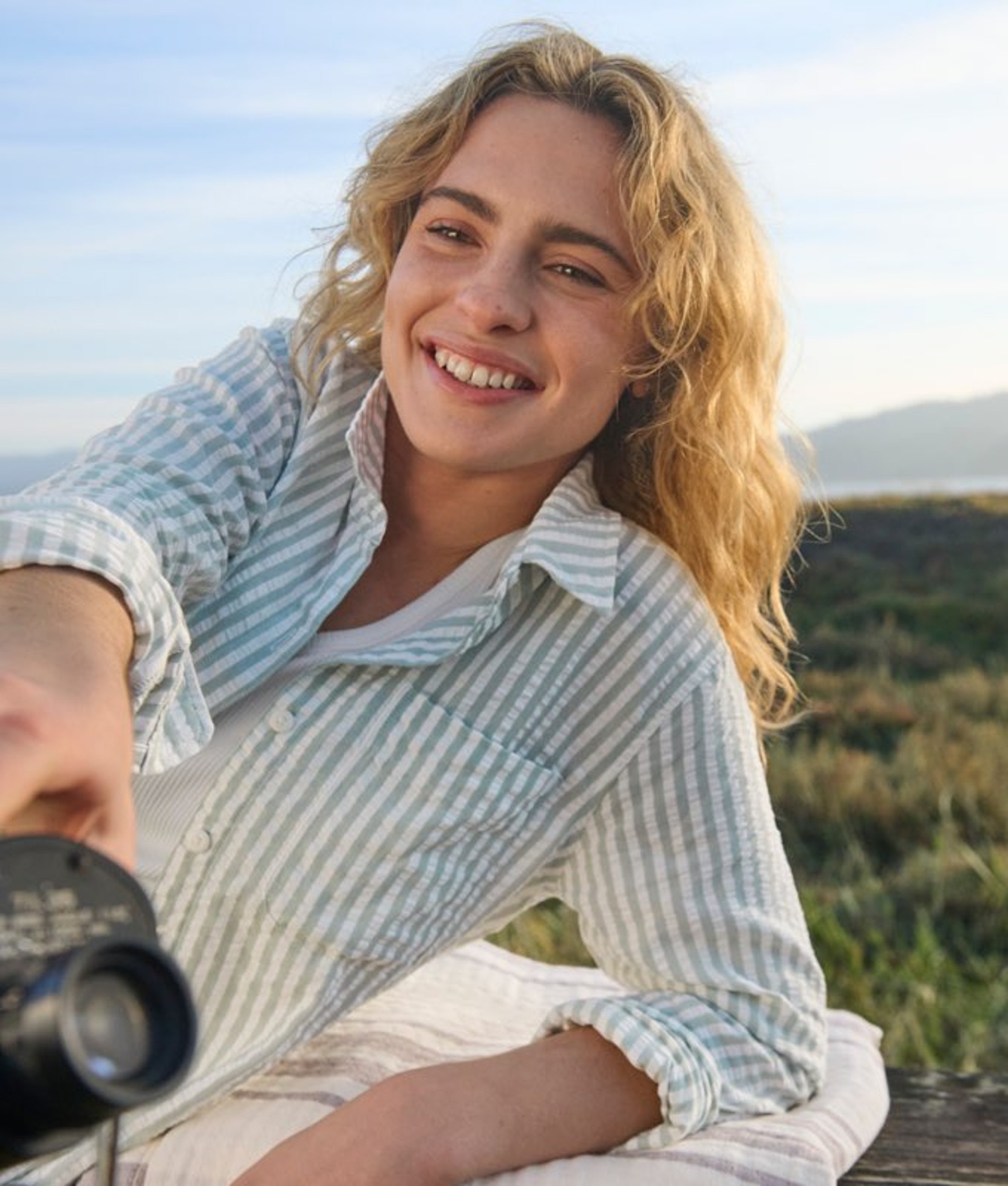Person wearing a striped short-sleeve shirt outdoors near the coast, with sunlight and water in the background.