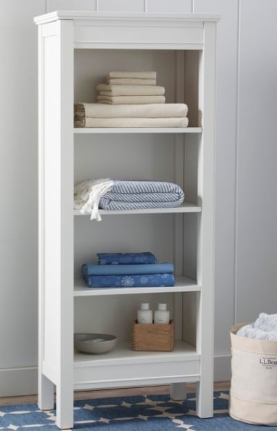 A tall white shelving unit with four shelves, holding neatly folded towels and toiletries. A basket with towels is on the floor beside it.