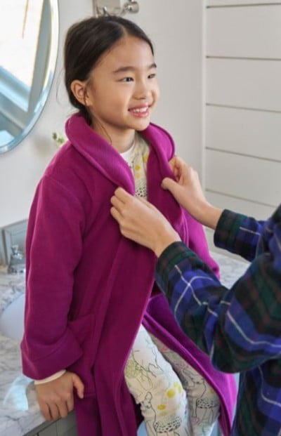 A child in printed pajamas sits on a bathroom counter while someone adjusts a bright magenta robe around them.