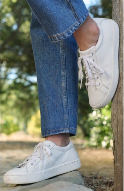 Woman wearing white sneakers and blue jeans stands outdoors with one foot on a wooden post.