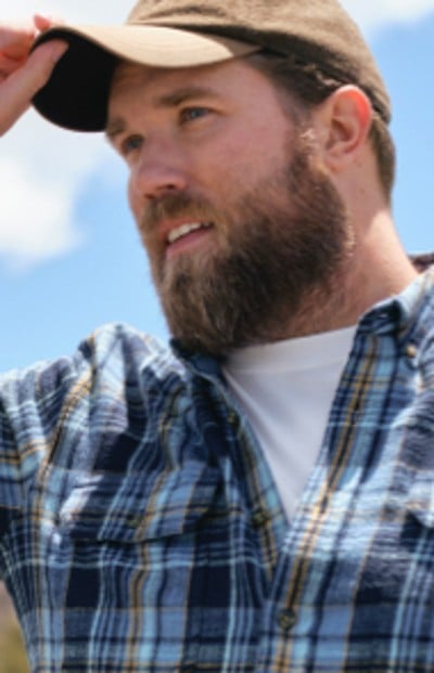 A man wearing a plaid shirt adjusts a baseball cap with a blue sky background.