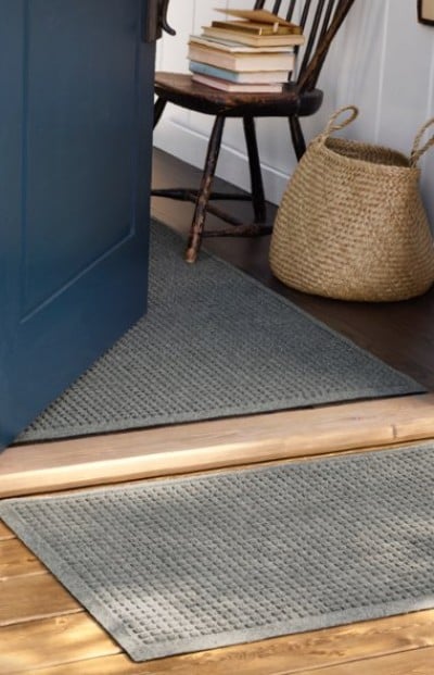Open blue door reveals a cozy entryway with gray rugs, a wooden chair stacked with books, and a woven basket on the floor.
