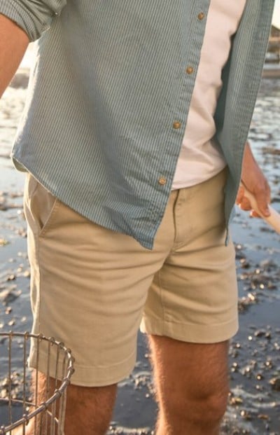 Person wearing a light blue button‑up shirt over a white tee with khaki shorts, standing near water.