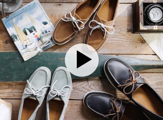 Top view of boat shoes arranged on a wooden surface with a magazine and compass with play video icon.