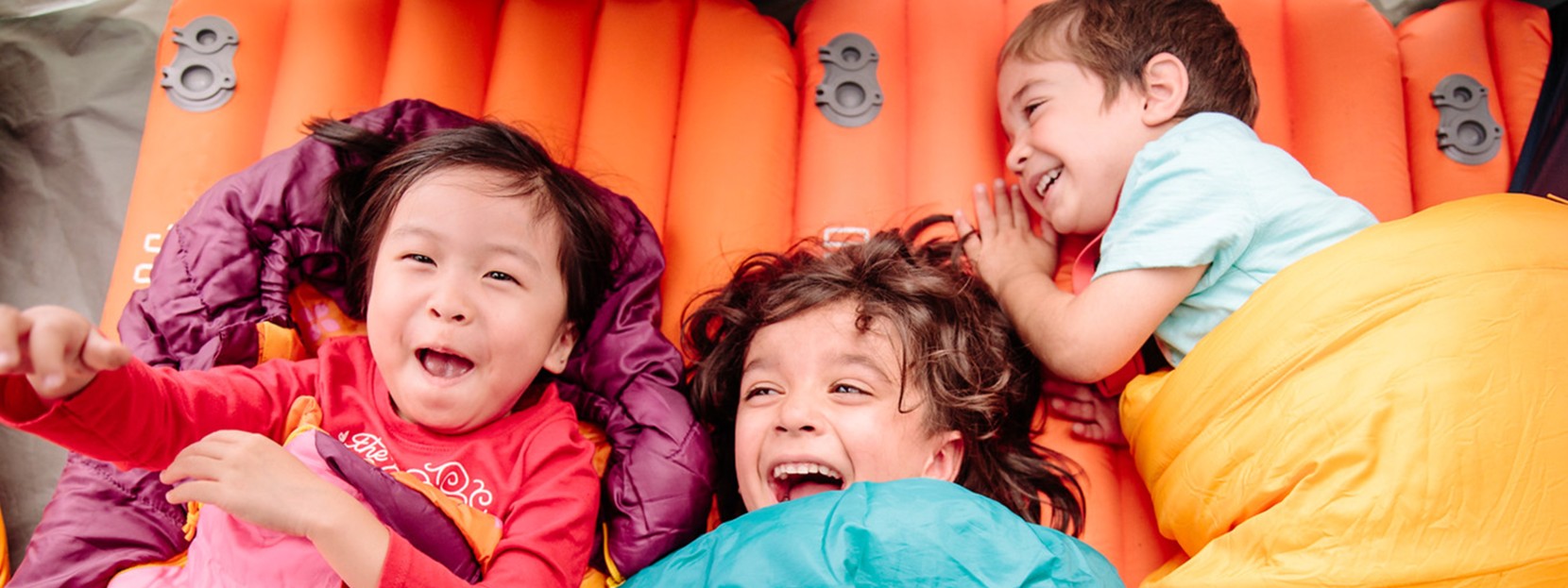 Three children laugh and lay in sleeping bags.