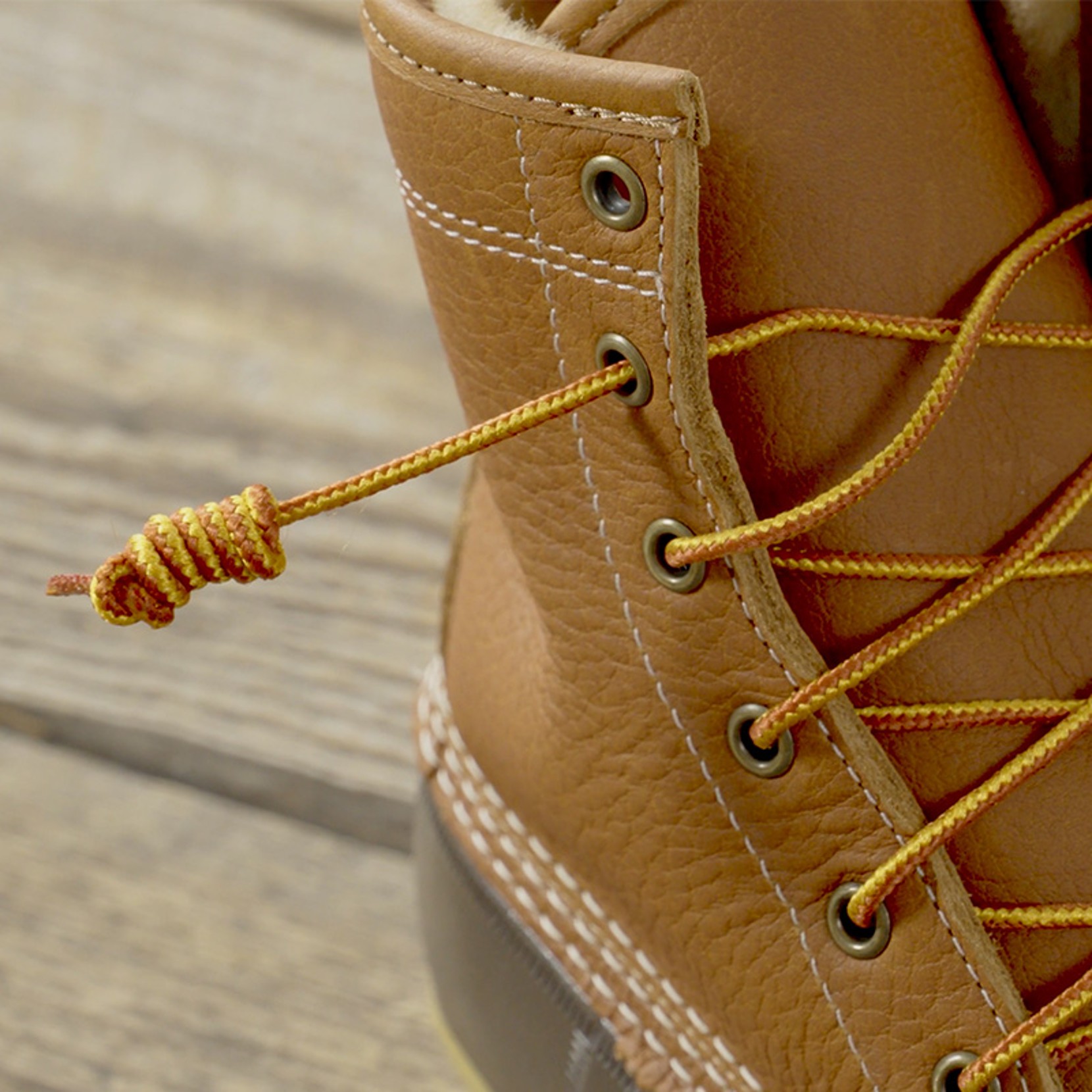 Close-up of brown leather boot with yellow laces, metal eyelets, and visible stitching on a wooden surface.