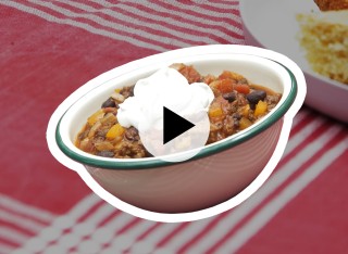A bowl of chili with a dollop of sour cream on a red and white checkered tablecloth with a play video icon in the center.