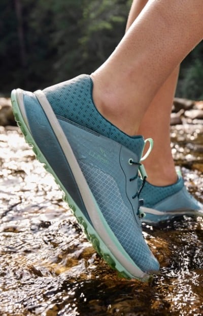 Person in blue athletic shoes stands in a shallow stream with water flowing around, surrounded by a forested background.