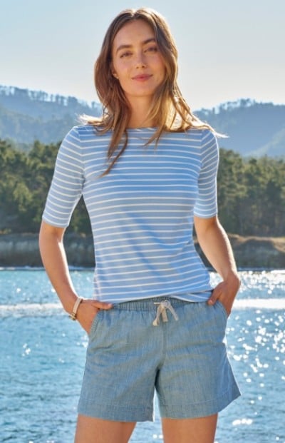 A woman wearing a blue and white striped shirt and blue shorts stands near water with hands in pockets. 