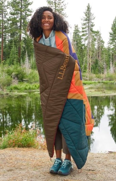 Wrapped in a colorful L.L.Bean outdoor blanket, a person stands near a forest pond wearing teal sneakers and cropped pants.