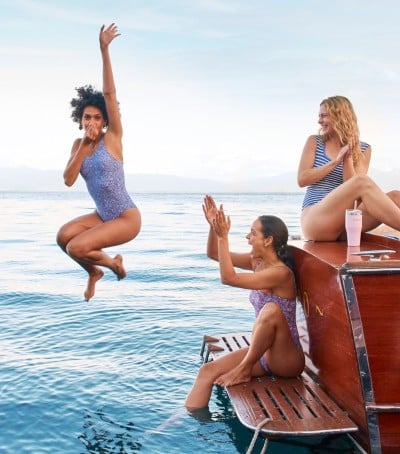 Person jumps from boat into lake as two others sit on the edge; clear blue water and sunny sky.