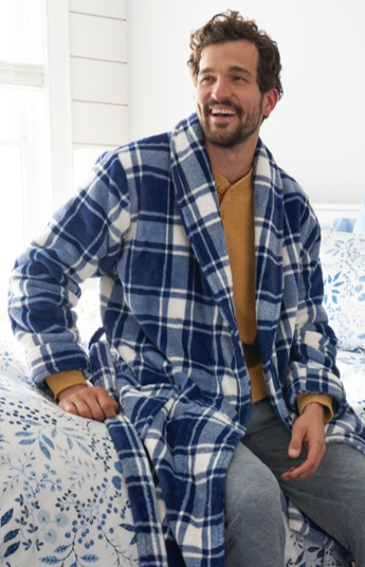 A man sits on a bed with a blue and white patterned bedspread, wearing a plaid robe over a yellow shirt and gray pants. 