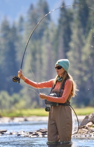 A person in waders and a blue beanie fly fishing in a river with a forested background.
