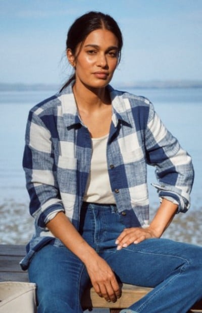Person sitting by the shore wearing a blue plaid shirt and jeans.