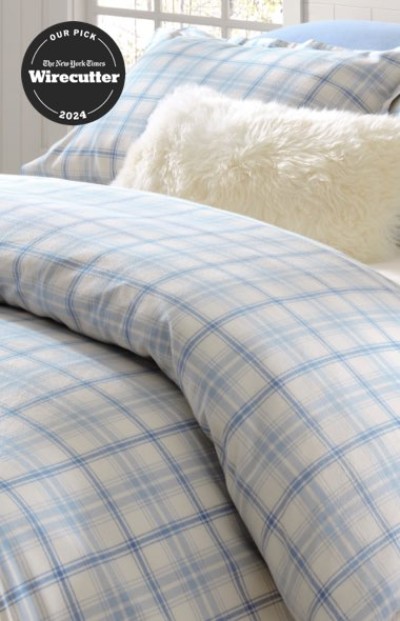 Soft blue plaid bedding with a fluffy white pillow on a bright, sunlit bed, New York Times Wirecutter logo in the corner.