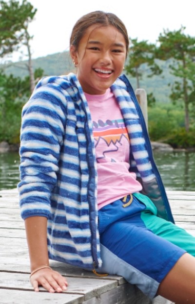 A girl sits outside on a wooden dock wearing a pink tshirt, striped hoodie and blue shorts.
