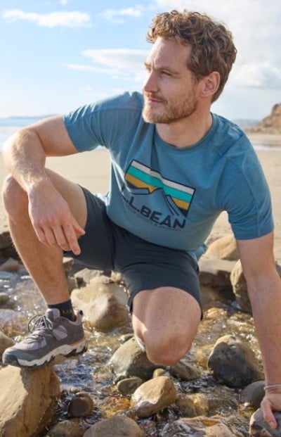 Person kneeling beside a small stream at the beach, wearing a blue L.L.Bean graphic T‑shirt, shorts, and hiking shoes.